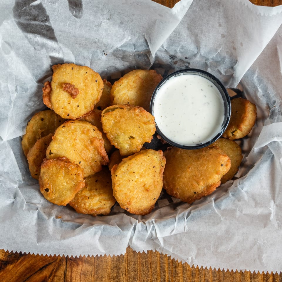 A basket of golden-fried pickles with a side of creamy ranch dressing on white parchment paper.