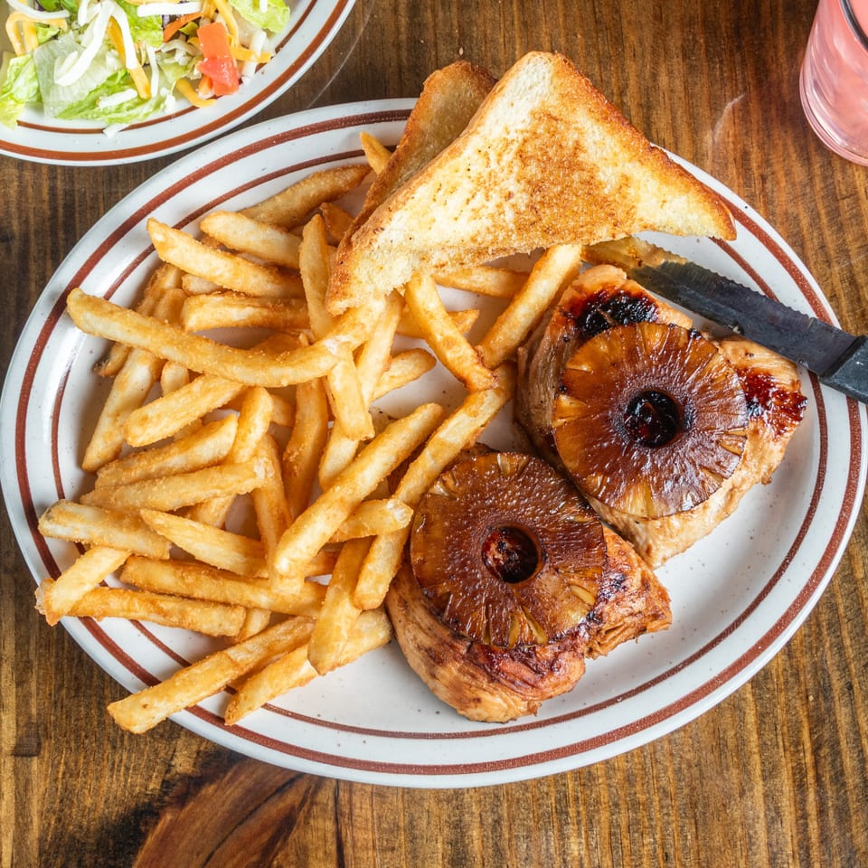 Grilled pineapple chicken with fries and toast on a white plate with brown stripes, next to a salad.