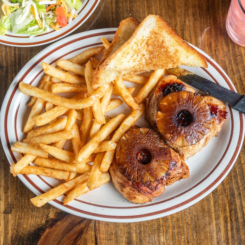 Grilled pineapple chicken with fries and toast on a white plate with brown stripes, next to a salad.
