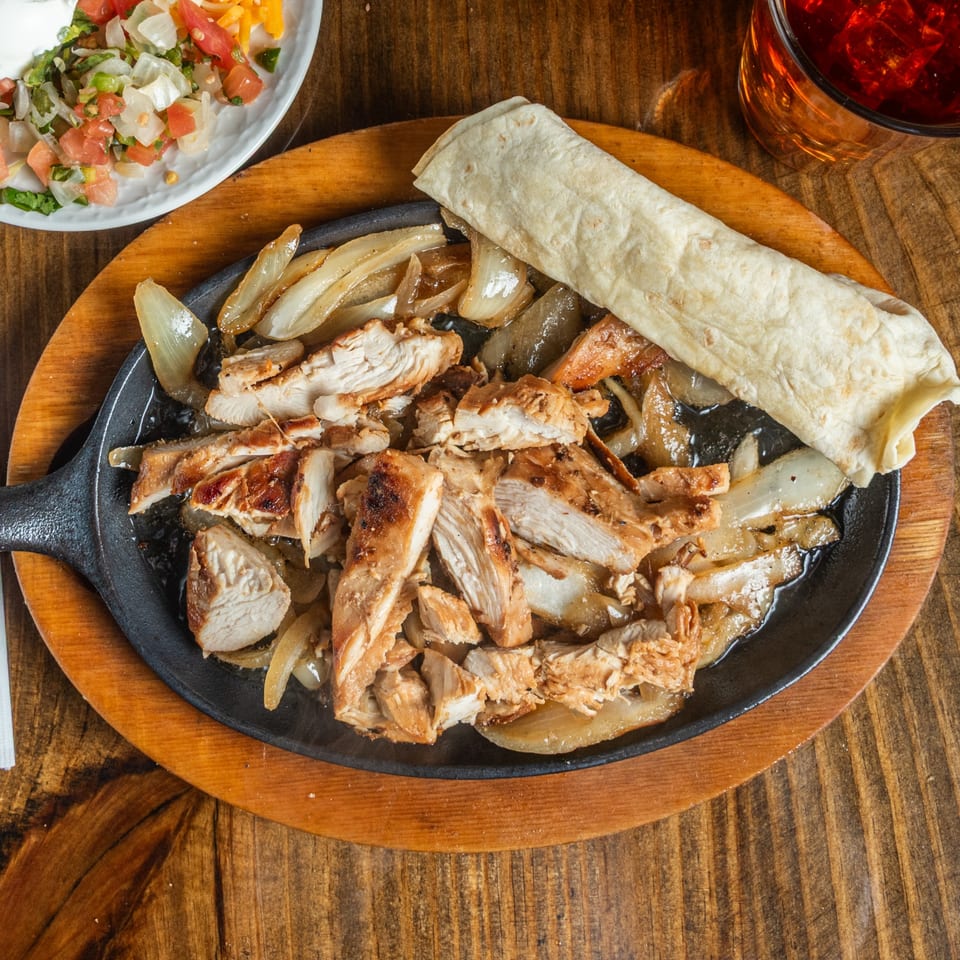 Sizzling chicken fajitas with onions, a tortilla, salsa, and a drink on a wooden table.