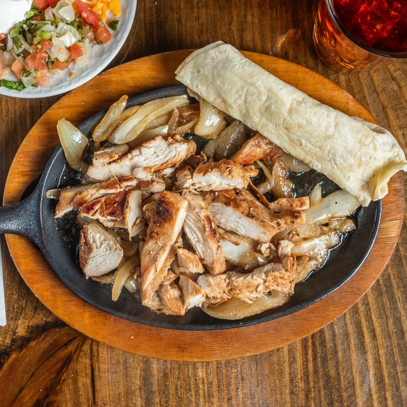 Sizzling chicken fajitas with onions, a tortilla, salsa, and a drink on a wooden table.