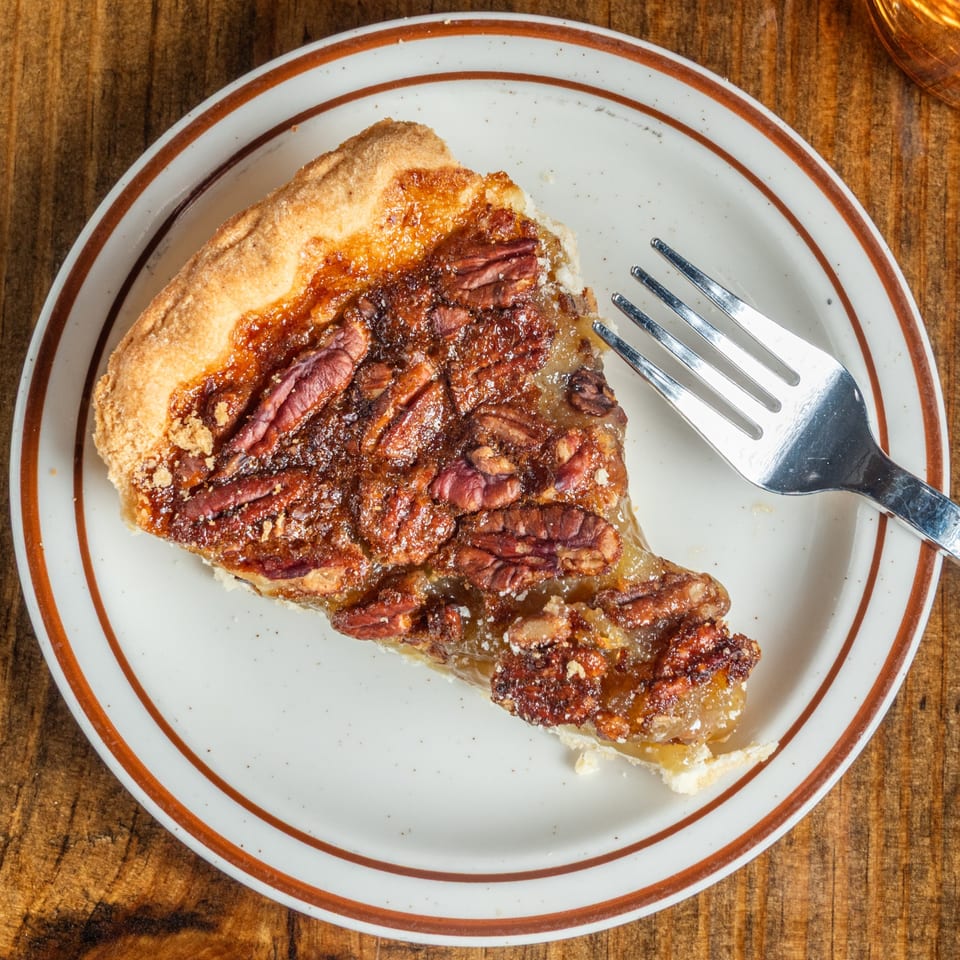 A slice of pecan pie with a fork on a white plate with brown stripes, on a wooden table.