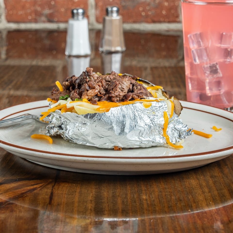 Baked potato with steak, cheese, and sour cream, wrapped in foil, with a pink drink.